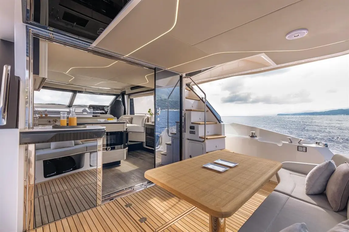 Modern yacht interior featuring a dining table, kitchenette, and large windows with ocean views. Drinks are visible on the counter.